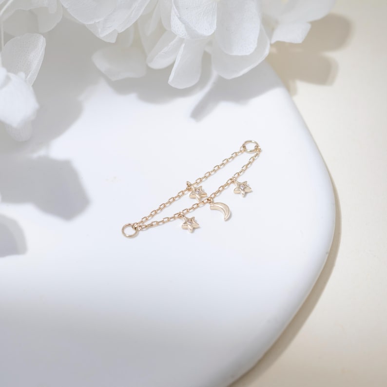 May include: A gold-toned double chain connector featuring two small star charms and one crescent moon charm. The delicate metal chain has a circular jump ring at each end, designed to link two points together with celestial-themed dangling accents.
