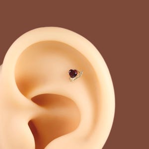 May include: A gold-toned ear stud featuring a small, faceted, deep red heart-shaped gemstone. The setting includes a delicate, curved metallic branch-like detail extending from the side of the heart, designed for placement in the ear cartilage.
