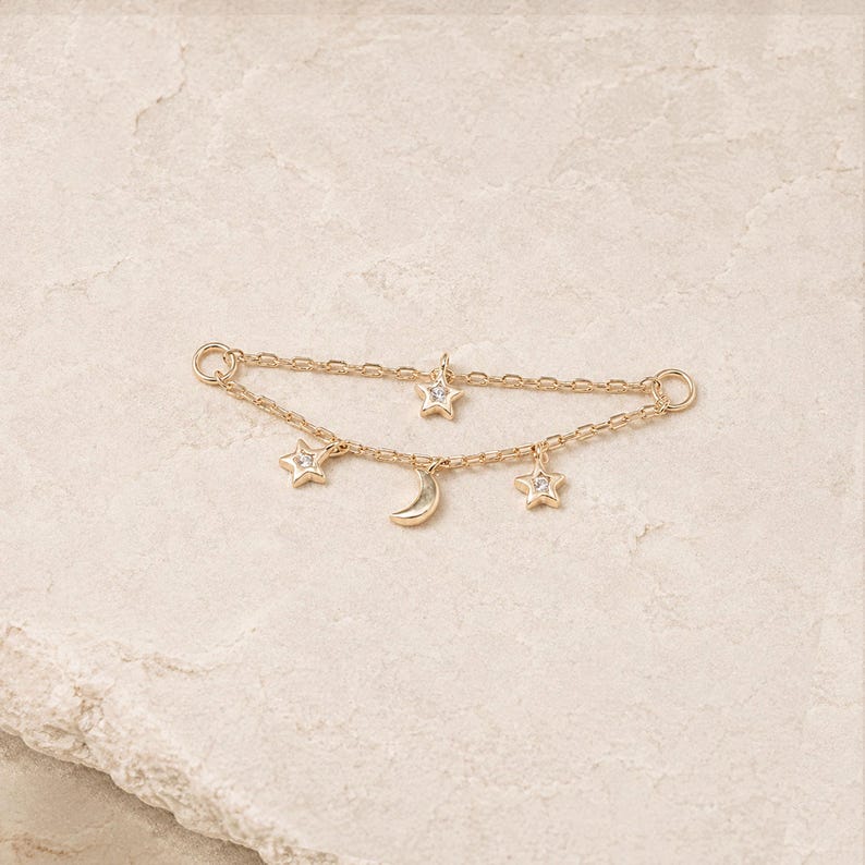 May include: A gold-toned jewelry connector chain featuring two delicate parallel chains. The top chain holds one small star charm, while the bottom chain features two star charms and a central crescent moon charm, all with clear stone accents.
