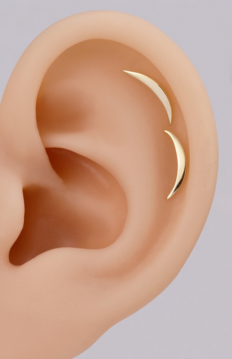 May include: Two gold-toned crescent moon-shaped earrings are positioned along the upper cartilage of an ear. Each earring features a smooth, polished surface with a curved, tapered design that follows the natural contour of the ear's outer rim.