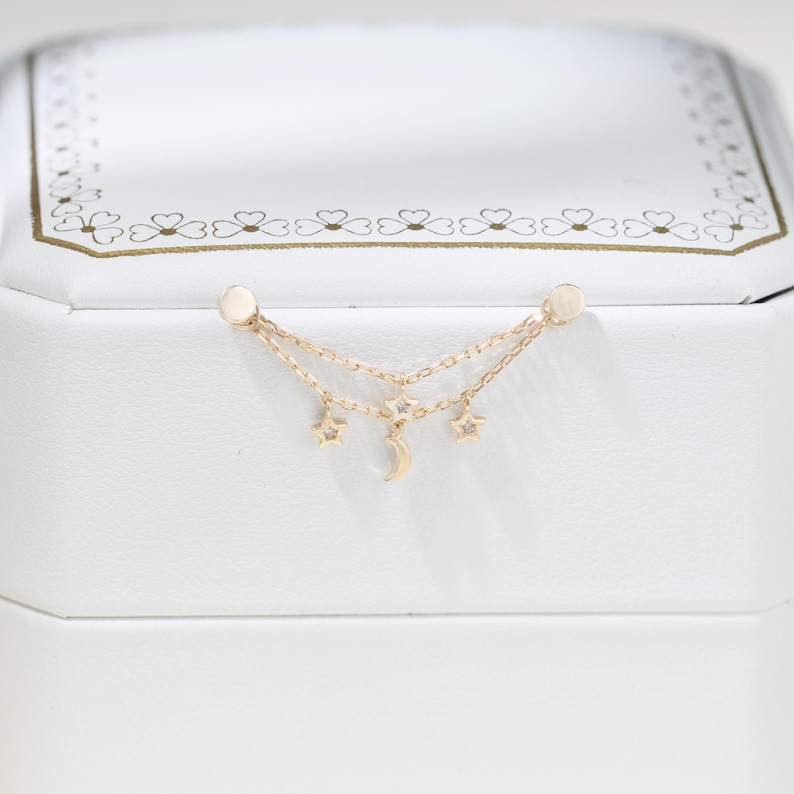 May include: A gold double-chain earring featuring two small star charms and a central crescent moon charm. Each star charm is accented with a tiny clear gemstone. The earring is designed with two flat circular posts at the ends of the chains.