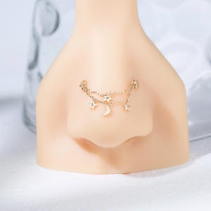 May include: A gold-toned nose chain featuring two delicate linked chains adorned with small star charms and a central crescent moon charm. The jewelry is attached to two circular studs, creating a layered effect across the bridge of the nose.