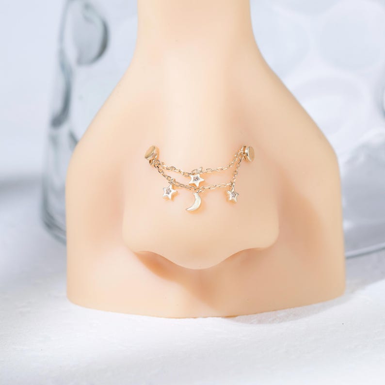May include: A gold-toned nose chain featuring two delicate linked chains adorned with small star charms and a central crescent moon charm. The jewelry is attached to two circular studs, creating a layered effect across the bridge of the nose.