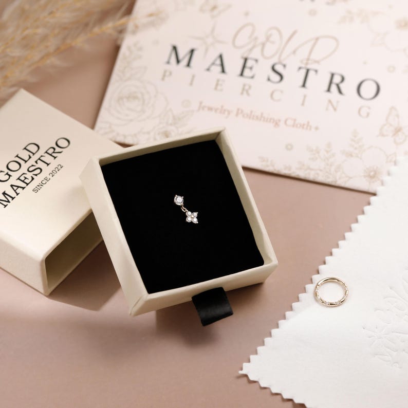 May include: A small gold piercing charm with two clear stones, one round and one flower-shaped, resting inside a cream-colored jewelry box with a black velvet insert. The box is labeled Gold Maestro, and a matching gold ring sits nearby.