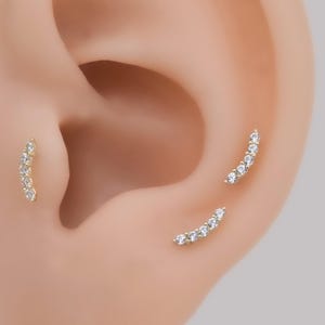 May include: Three curved ear piercings featuring a gold-toned metal bar set with five small, clear, round-cut stones. Each piece follows a gentle arc, designed to sit along the cartilage or lobe, providing a delicate and shimmering appearance.