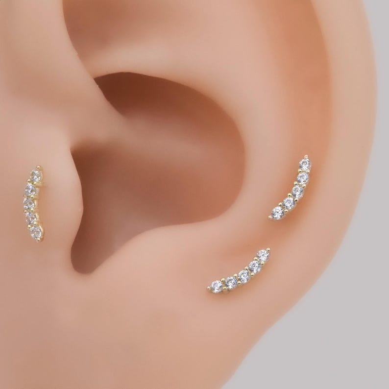 May include: Three curved ear piercings featuring a gold-toned metal bar set with five small, clear, round-cut stones. Each piece follows a gentle arc, designed to sit along the cartilage or lobe, providing a delicate and shimmering appearance.