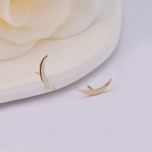 May include: A pair of gold-toned stud earrings shaped like crescent moons. Each earring features a smooth, polished surface and a thin post extending from the back, designed to be worn through the earlobe with a simple, curved silhouette.
