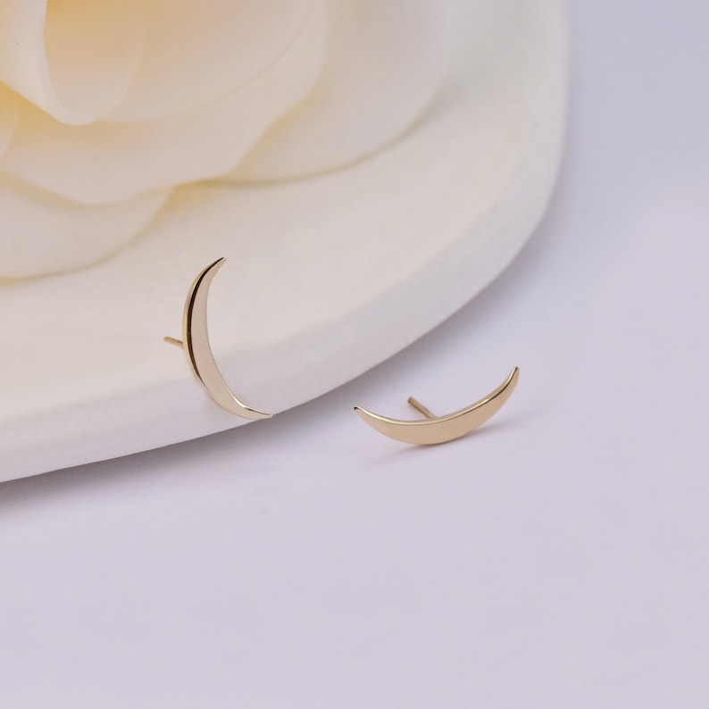 May include: A pair of gold-toned stud earrings shaped like crescent moons. Each earring features a smooth, polished surface and a thin post extending from the back, designed to be worn through the earlobe with a simple, curved silhouette.