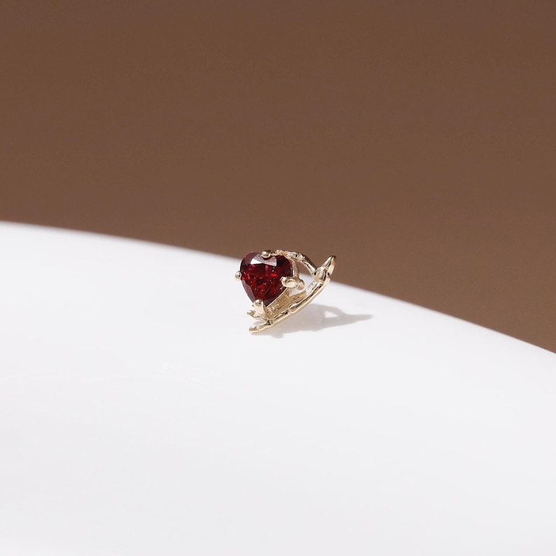 May include: A gold-toned stud earring featuring a deep red, heart-shaped gemstone held by a delicate, textured metal setting that curves around the base of the stone, creating a small, intricate piece of jewelry for the ear.