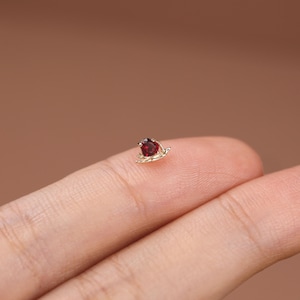 May include: A small gold stud earring featuring a heart-shaped red gemstone set in a delicate metal frame. The piece is crafted with a polished gold finish and a prong setting that secures the deep red stone at the center of the heart design.