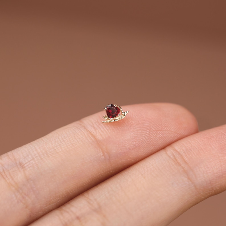 May include: A small gold stud earring featuring a heart-shaped red gemstone set in a delicate metal frame. The piece is crafted with a polished gold finish and a prong setting that secures the deep red stone at the center of the heart design.