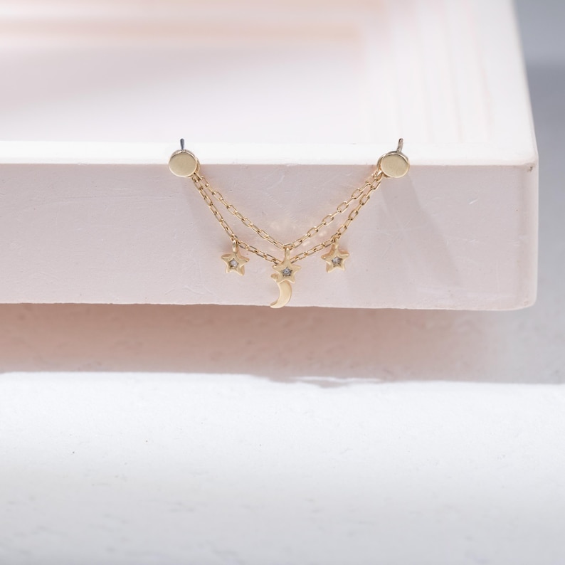 May include: A gold-toned double chain earring featuring two circular studs connected by two delicate chains. The chains are adorned with two small star charms and a central crescent moon charm, each accented with a tiny embedded clear stone.