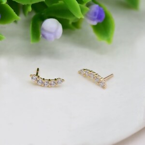 May include: A pair of gold stud earrings, each featuring a curved bar design set with five small, clear, round-cut stones. The earrings have a polished gold finish and a standard post backing, showcasing a delicate and symmetrical arrangement.