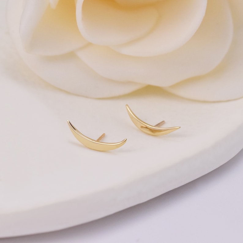 May include: A pair of gold-toned stud earrings shaped like slender, curved crescent moons. Each earring features a smooth, polished metallic surface with a pointed post extending from the back, designed to sit flush against the earlobe when worn.
