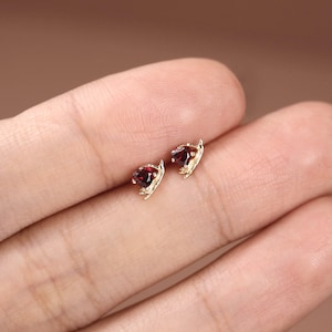 May include: A pair of small gold stud earrings, each featuring a faceted, deep red garnet gemstone set in a delicate, leaf-inspired metal frame that curves around the base of the stone, creating a subtle, organic, and refined appearance.