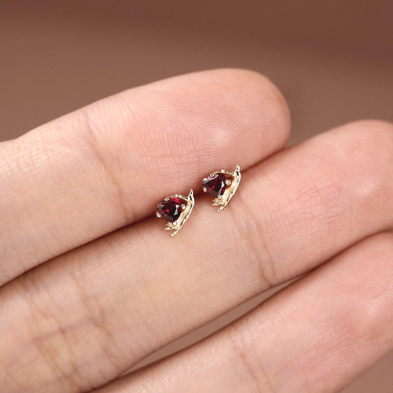 May include: A pair of small gold stud earrings, each featuring a faceted, deep red garnet gemstone set in a delicate, leaf-inspired metal frame that curves around the base of the stone, creating a subtle, organic, and refined appearance.