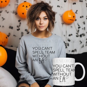 Peut inclure: Une tasse en céramique blanche avec un motif noir qui dit "You can't spell team without an E.A." avec un petit symbole de cœur. La tasse est posée sur une table à côté d'une personne portant un sweat-shirt gris avec le même motif.