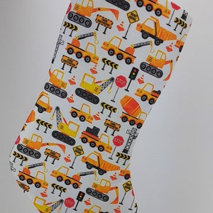 May include: A white Christmas stocking with a construction theme. The stocking is covered in a repeating pattern of yellow construction vehicles, traffic cones, and road signs. The signs say "Road Work", "Under Construction", and "Stop".