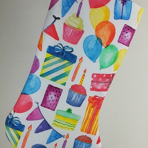 May include: A white stocking with a birthday theme. The stocking is decorated with colorful watercolor illustrations of cupcakes, presents, balloons, and flags.