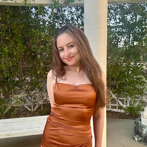 May include: A sleeveless, copper-colored satin dress featuring a straight neckline, thin spaghetti straps, and a fitted bodice that transitions into a gathered, ruched skirt detail along the side, creating a fluid and draped silhouette.