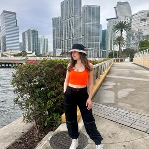 May include: A person wearing a black bucket hat with a colorful pattern, an orange crop top, black cargo pants, and white sneakers. The person is standing on a sidewalk in front of a city skyline.