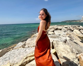 Könnte beinhalten: Eine Frau in einem rostfarbenen Maxikleid steht an einem felsigen Ufer mit dem Meer im Hintergrund. Der Himmel ist blau und es gibt weiße Wolken.