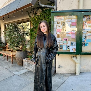 Peut inclure: Un long trench-coat noir avec une ceinture, porté à l'extérieur. Le manteau a un aspect brillant et texturé. La personne porte des bottes noires et un haut noir. L'arrière-plan comprend un bâtiment avec un tableau d'affichage vert et un banc.