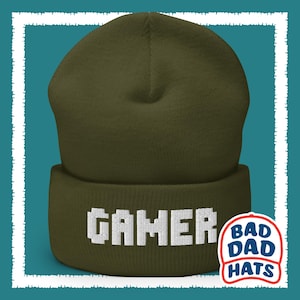May include: A green beanie with the word "GAMER" embroidered in white on the front. The beanie has a white logo on the side that says "BAD DAD HATS".