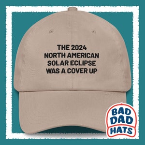 May include: A tan baseball cap with black embroidered text that reads "THE 2024 NORTH AMERICAN SOLAR ECLIPSE WAS A COVER UP". The cap has a black and white logo on the back that reads "BAD DAD HATS".