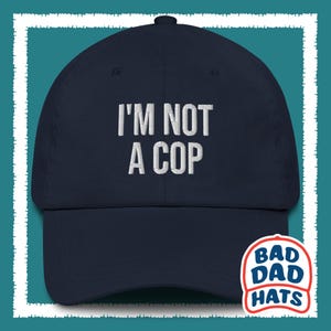 Può includere: Un cappello da baseball blu navy con ricamo bianco che recita "I'M NOT A COP". Il cappello ha una visiera curva e un logo bianco sul retro che recita "BAD DAD HATS".