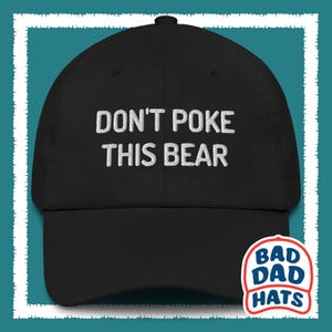 May include: A black baseball cap featuring the white embroidered text DON'T POKE THIS BEAR centered on the front panel. The hat has a curved brim and a structured crown. A small logo with the text BAD DAD HATS is visible in the bottom corner.