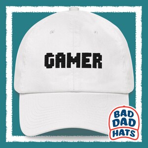 May include: White baseball cap with black pixelated text that reads "GAMER". The cap has a curved brim and a Bad Dad Hats logo on the side.