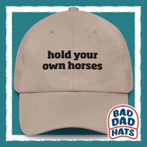 May include: A tan baseball cap with black embroidery that reads "hold your own horses". The cap has a black and red logo on the side that reads "Bad Dad Hats".