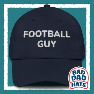 May include: A navy blue baseball cap with white embroidered text that reads "FOOTBALL GUY". The cap has a curved brim and a white logo on the back that reads "BAD DAD HATS".