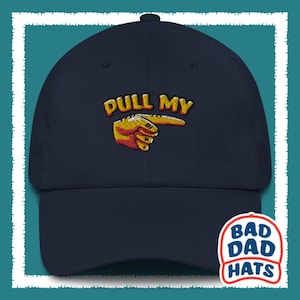 May include: A navy blue baseball cap with the text "PULL MY" embroidered in yellow and red. A yellow hand with a red finger pointing to the right is embroidered below the text. The cap has a white label with the text "BAD DAD HATS" on the side.