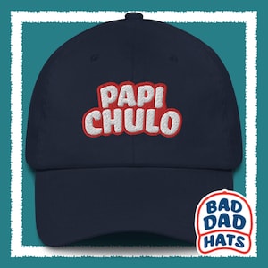 May include: A navy blue baseball cap with a red and white embroidered logo that says "Papi Chulo". The logo has a white outline and a 3D effect. The cap has a white adjustable strap and a white label that says "Bad Dad Hats" on the back.