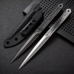 May include: Two silver and black daggers with pointed blades and patterned handles are displayed. A black sheath with a clip is next to the daggers. The daggers are on a dark surface.