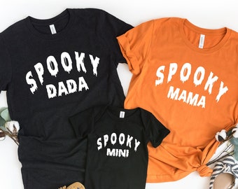 May include: Three black and orange t-shirts with the text "Spooky Dada", "Spooky Mama", and "Spooky Mini" in white dripping letters. The shirts are laid out on a white background with cotton stems and black and white striped shoes.