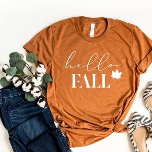 May include: A rust-colored t-shirt with the text "hello fall" in white with a white leaf graphic. The shirt is laid out on a white surface with a pair of black and white striped shoes, a pair of blue jeans, a sprig of eucalyptus, and two small pumpkins.