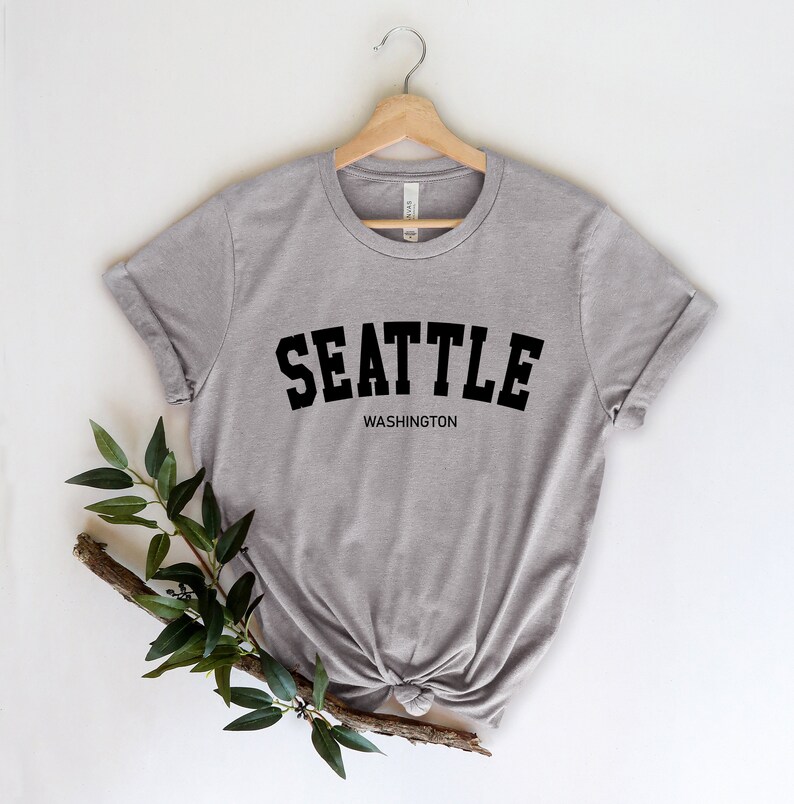 Peut inclure: T-shirt gris chin&eacute; avec le mot "SEATTLE" en grosses lettres noires arqu&eacute;es, et "WASHINGTON" en dessous. Le t-shirt est suspendu &agrave; un cintre en bois sur fond blanc.