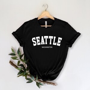 Peut inclure: T-shirt noir avec le mot "SEATTLE" en grandes lettres blanches au-dessus de "WASHINGTON" en plus petites lettres blanches. Le t-shirt est suspendu &agrave; un cintre en bois sur fond blanc.