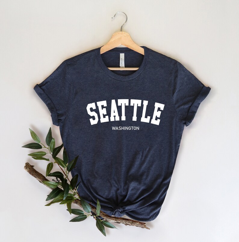 Peut inclure: T-shirt bleu marine fonc&eacute; avec le mot "SEATTLE" en lettres blanches en gras au-dessus de "WASHINGTON". Le t-shirt est pr&eacute;sent&eacute; sur un cintre en bois avec une branche de verdure.