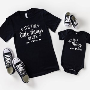 May include: A black t-shirt with the text "It's the little things in life" and a white arrow with a heart in the middle. A black bodysuit with the text "little thing" and a white arrow with a heart in the middle. Both shirts have a white graphic design.
