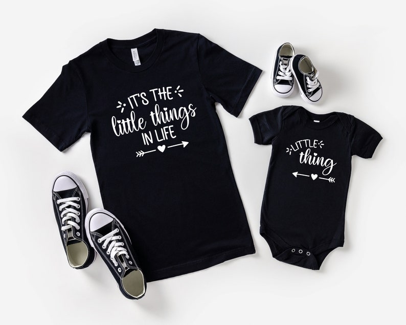 May include: A black t-shirt with the text "It's the little things in life" and a white arrow with a heart in the middle. A black bodysuit with the text "little thing" and a white arrow with a heart in the middle. Both shirts have a white graphic design.