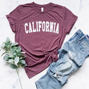 K&ouml;nnte beinhalten: Ein maroonfarbenes T-Shirt mit dem Wort "CALIFORNIA" in wei&szlig; auf der Vorderseite. Das Shirt liegt flach auf einer wei&szlig;en Marmorfl&auml;che mit einem Zweig Gr&uuml;n und einer hellblauen Jeans mit Destroyed-Details.