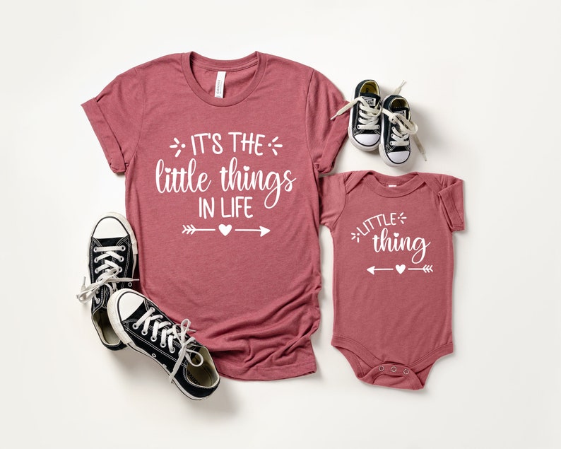 May include: A set of two shirts with a matching design. The larger shirt is a maroon t-shirt with the text "It's the little things in life" and two arrows with a heart in the middle. The smaller shirt is a maroon bodysuit with the text "little thing" and two arrows with a heart in the middle.