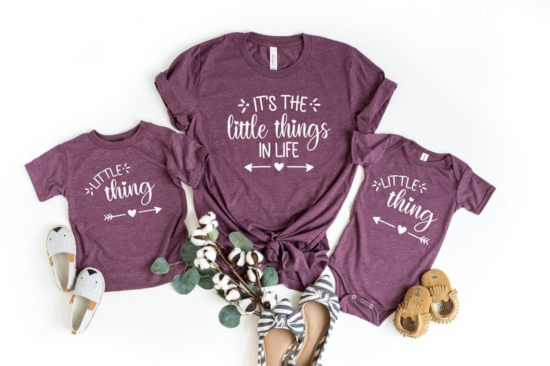 May include: Three maroon t-shirts with white text. The largest shirt says "It's the little things in life" with arrows and a heart. The two smaller shirts say "little thing" with an arrow and a heart.