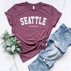 Peut inclure: Un t-shirt bordeaux avec le mot "SEATTLE" en lettres blanches, avec "WASHINGTON" en dessous. Il est associ&eacute; &agrave; un jean bleu d&eacute;lav&eacute; et une branche de feuilles vertes, le tout sur une surface en marbre blanc.