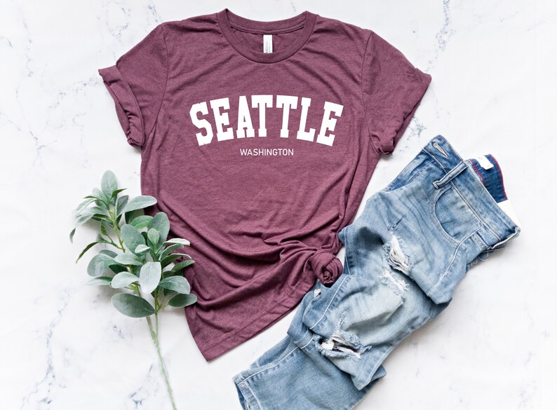Peut inclure: Un t-shirt bordeaux avec le mot "SEATTLE" en lettres blanches, avec "WASHINGTON" en dessous. Il est associ&eacute; &agrave; un jean bleu d&eacute;lav&eacute; et une branche de feuilles vertes, le tout sur une surface en marbre blanc.