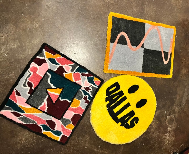May include: Three colorful rugs with geometric and abstract designs. One rug is a yellow circle with a black smiley face and the text "DALLAS" inside. Another rug is a square with a black and white checkered pattern and a pink wavy line. The third rug is a square with a multi-colored abstract design.